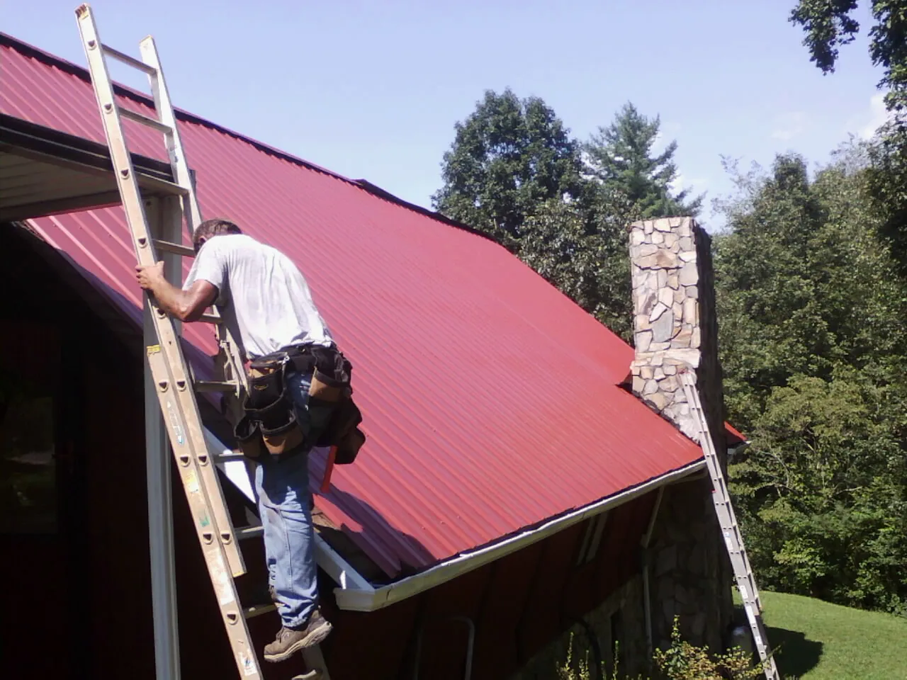 Roofer on a ladder beside a red metal roof and stone chimney — Soffit & Fascia Repair by Roofing Brothers in Arthur